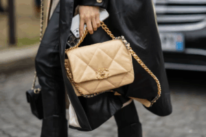 Close-up of a beige Chanel quilted handbag with gold chain strap, carried by a person wearing a black trench coat and holding a phone.