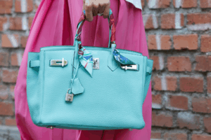 A vibrant turquoise Birkin with twilly-wrapped handles adds a pop of color against a brick wall backdrop.