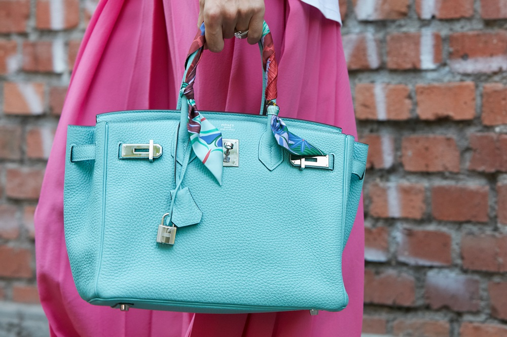 A vibrant turquoise Birkin with twilly-wrapped handles adds a pop of color against a brick wall backdrop. A vibrant turquoise Birkin with twilly-wrapped handles adds a pop of color against a brick wall backdrop.