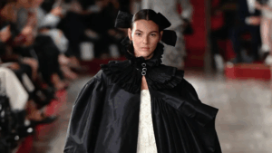 Model in a dramatic black cape with ruffled high collar and oversized bows walking down the runway.