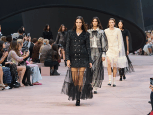 Models walk the Chanel runway wearing black and white ensembles featuring tweed and sheer tulle overlays, while the audience captures the moment on their phones.