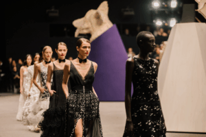 Models in monochrome black and white gowns walk the runway at a fashion show, featuring dramatic silhouettes, floral appliqués, and sharp bow-tie chokers.