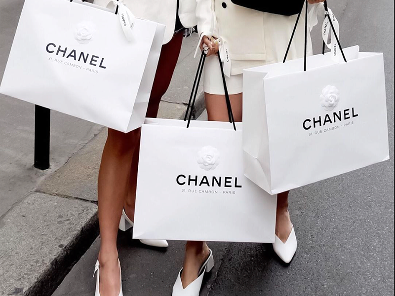 Group of people standing on a sidewalk, carrying several large white Chanel shopping bags with black logo and handles.