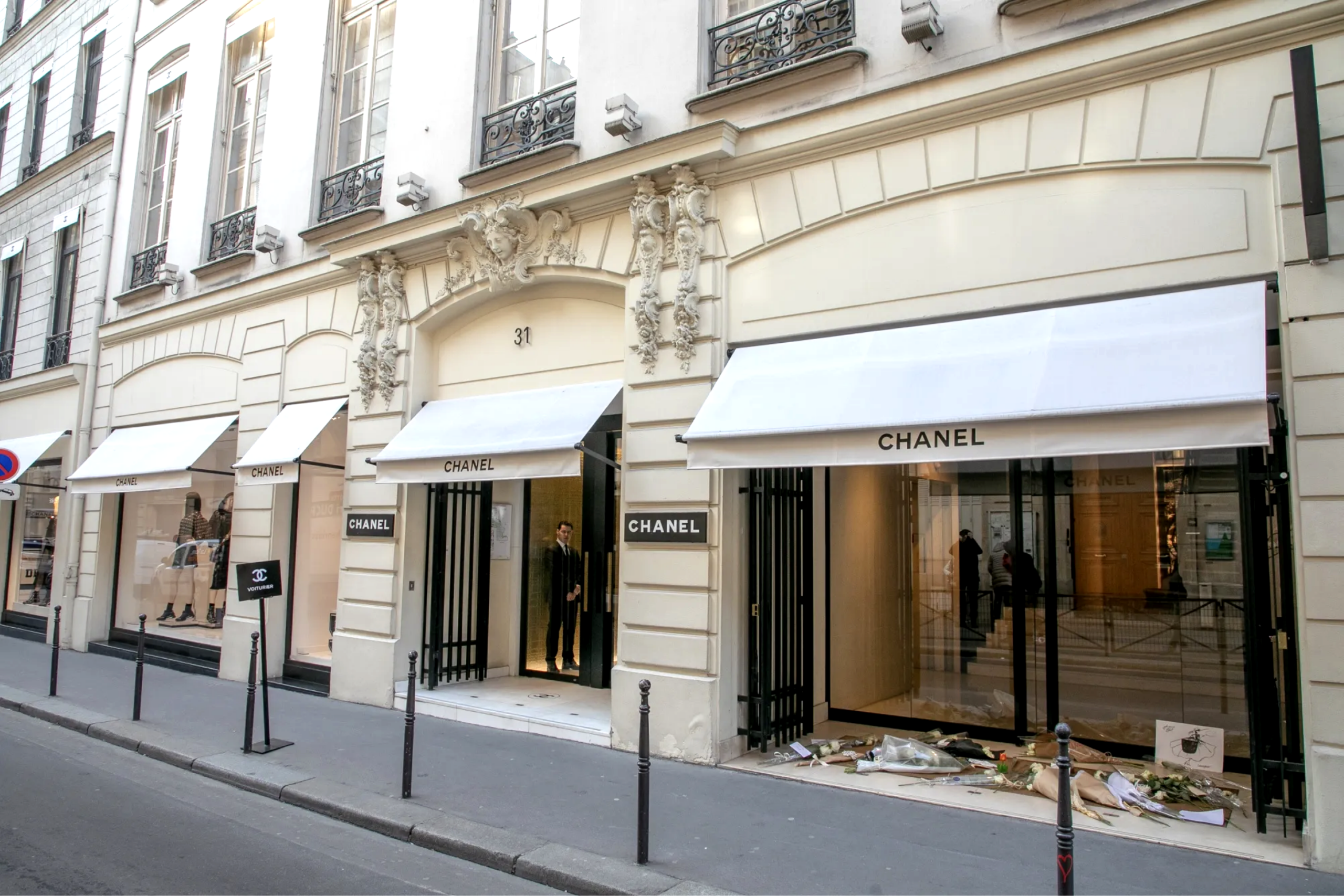 Chanel storefront on a Parisian street with white awnings, large glass doors, and ornate stone sculptures above the entrance.