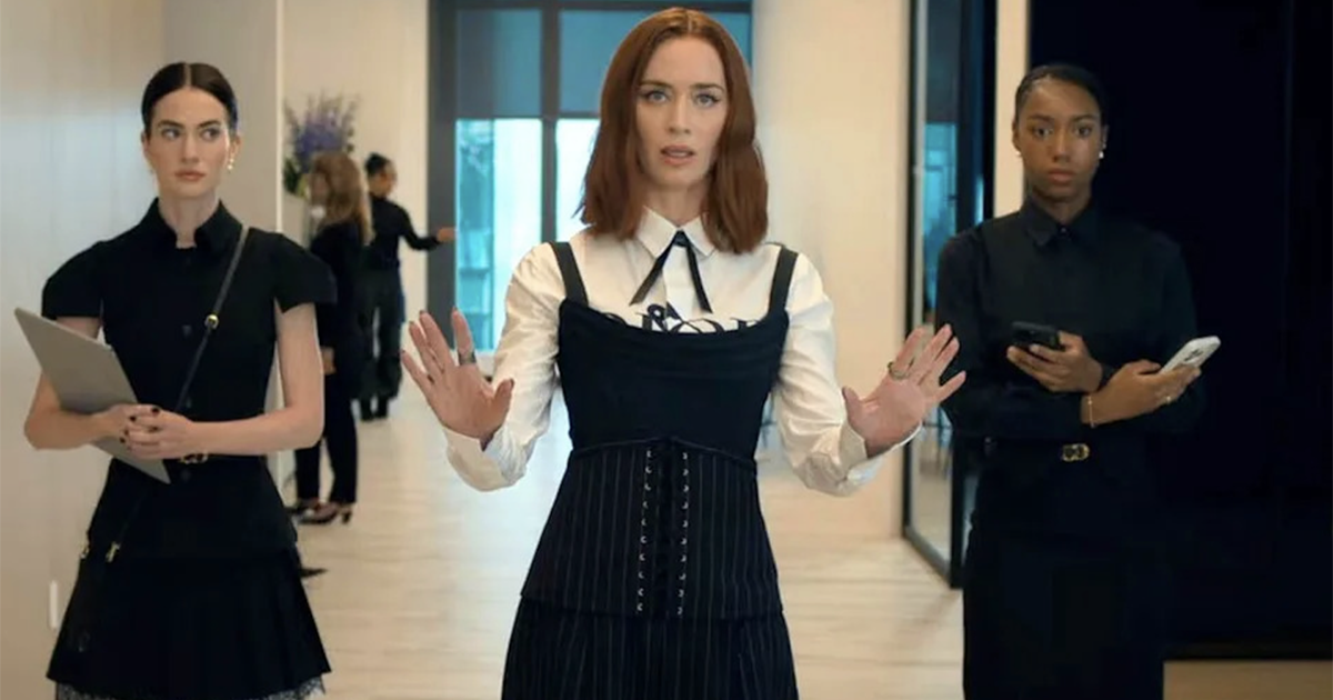 Center woman in a white shirt and black corset-style dress gestures with hands raised as two colleagues flank her in a modern office hallway with glass walls on the right and a folder in the left colleague's arms for context.