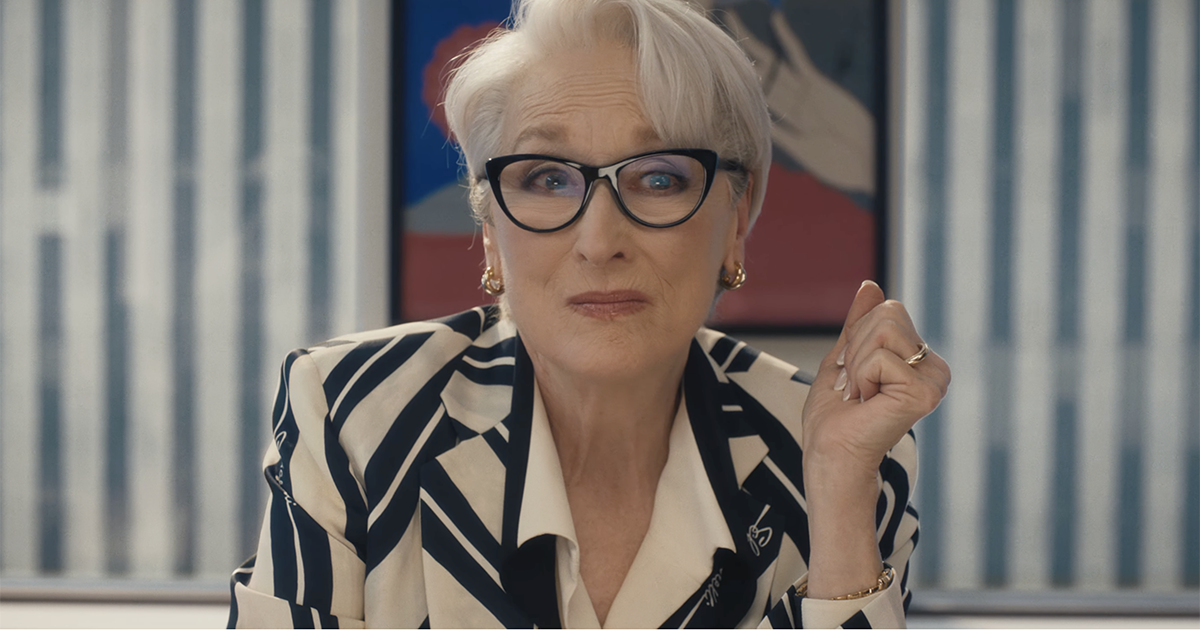 Older woman with short white hair and black cat-eye glasses sits at a desk, looking thoughtful with a slight smile.
